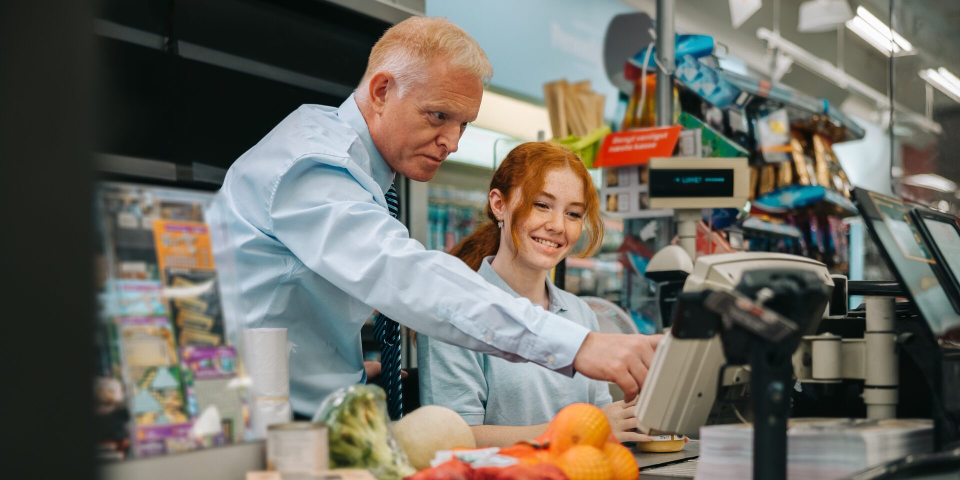 Senior manager helping new employee at checkout counter in supermarket. Mature man showing and explaining the checkout system to a trainee.