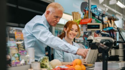 Senior manager helping new employee at checkout counter in supermarket. Mature man showing and explaining the checkout system to a trainee.